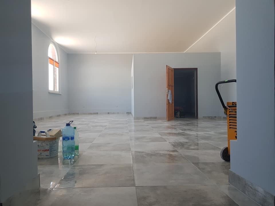 Wnętrze po malowaniu na szaro, widoczne materiały malarskie, marmurowe płytki podłogowe i wózek transportowy.