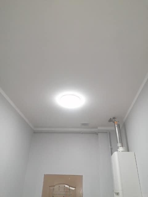 Pomieszczenie po remoncie, widoczne gładkie, białe ściany i sufit z okrągłym oświetleniem wpuszczanym, rury grzewcze pod sufitem oraz piec gazowy w rogu.