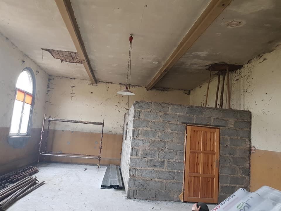 Wnętrze remontowanego pomieszczenia z widocznym fragmentem uszkodzonego sufitu, rusztowaniem, ścianą z pustaków z drewnianymi drzwiami i materiałami budowlanymi na podłodze.