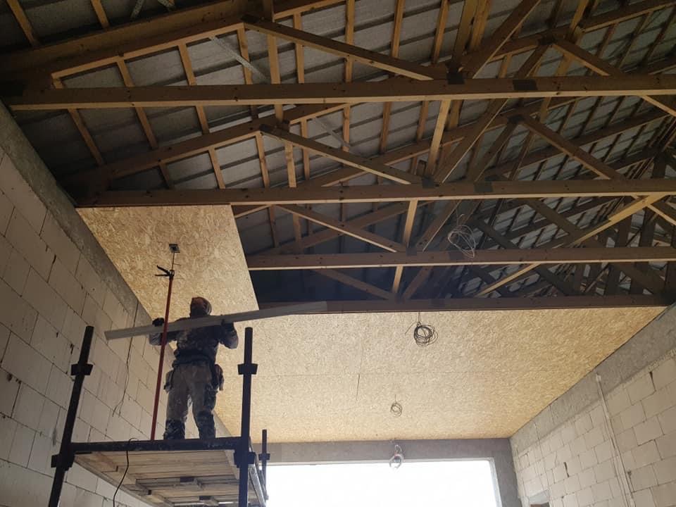 Pracownik na rusztowaniu montuje płytę OSB na suficie poddasza, widoczne drewniane elementy konstrukcji dachu i surowy mur