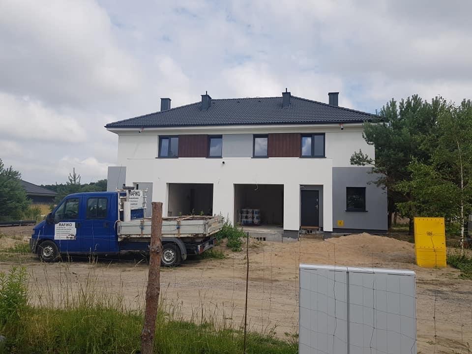 Dwukondygnacyjny dom w trakcie malowania elewacji, z widocznym firmowym samochodem dostawczym i materiałami budowlanymi na posesji.