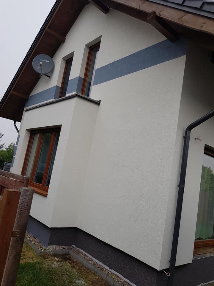 Wykończona elewacja domu w Gnieźnie z białym tynkiem i szarym pasem dekoracyjnym, widoczna antena satelitarna i drewniane okna.
