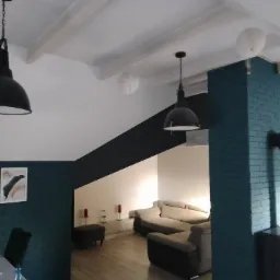 Salon z pomalowaną na turkusowo cegłą, czarnym kominem i lampami w stylu industrialnym oraz drewnianymi belkami stropowymi.