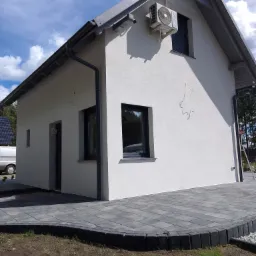 Wykończona elewacja domu jednorodzinnego z klimatyzatorem, widoczne ślady po instalacji elektrycznej, szara kostka brukowa wokół budynku.