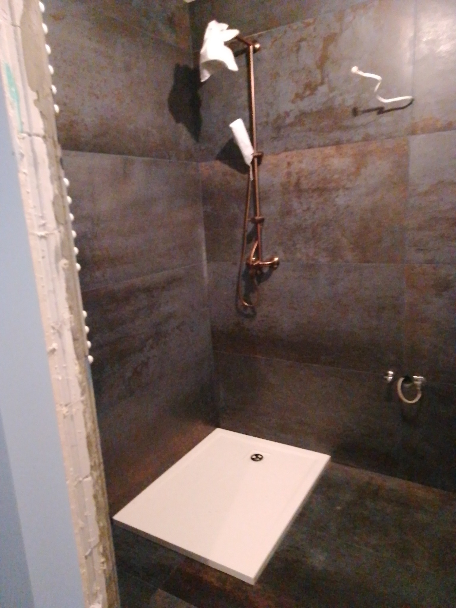 Nowoczesny brodzik w łazience z ciemnymi, metalicznymi płytkami na ścianach i miedzianą armaturą prysznicową. Widoczne ślady prac wykończeniowych przy ścianie.