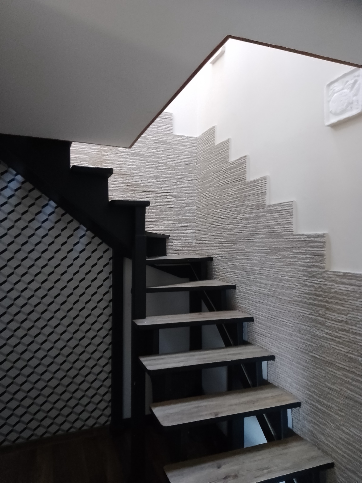 Nowoczesne schody z drewnianymi stopniami i czarną metalową konstrukcją, ściana wykończona dekoracyjną szarą cegłą, obok ściana z geometryczną tapetą.