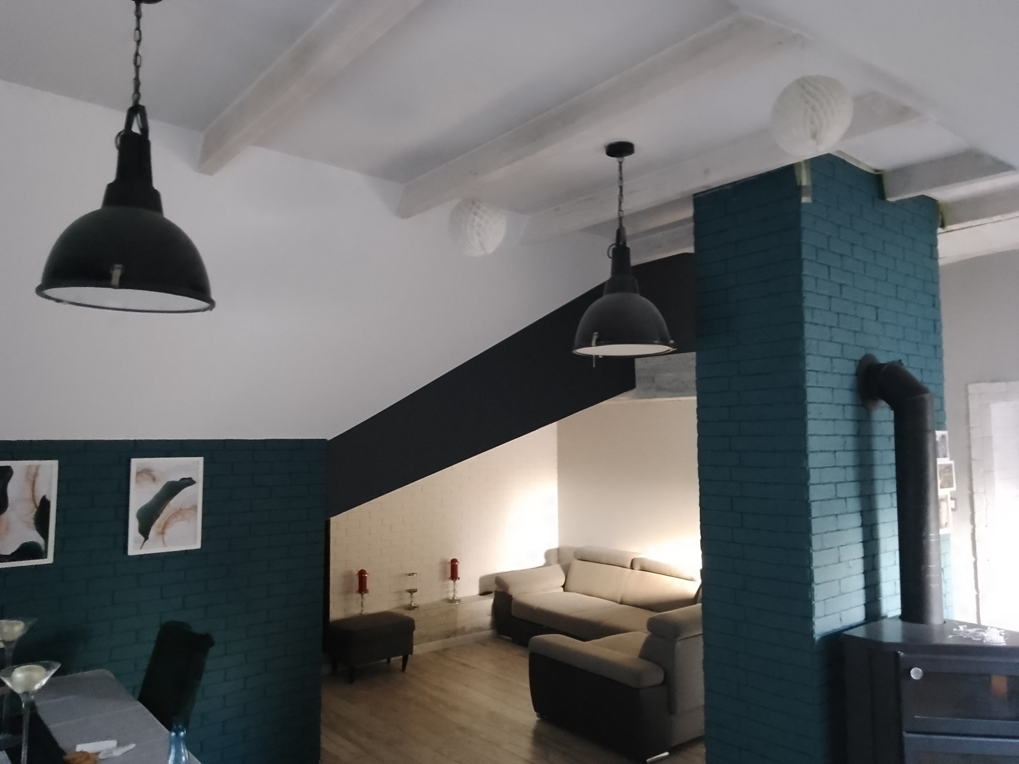 Salon z pomalowaną na turkusowo cegłą, czarnym kominem i lampami w stylu industrialnym oraz drewnianymi belkami stropowymi.