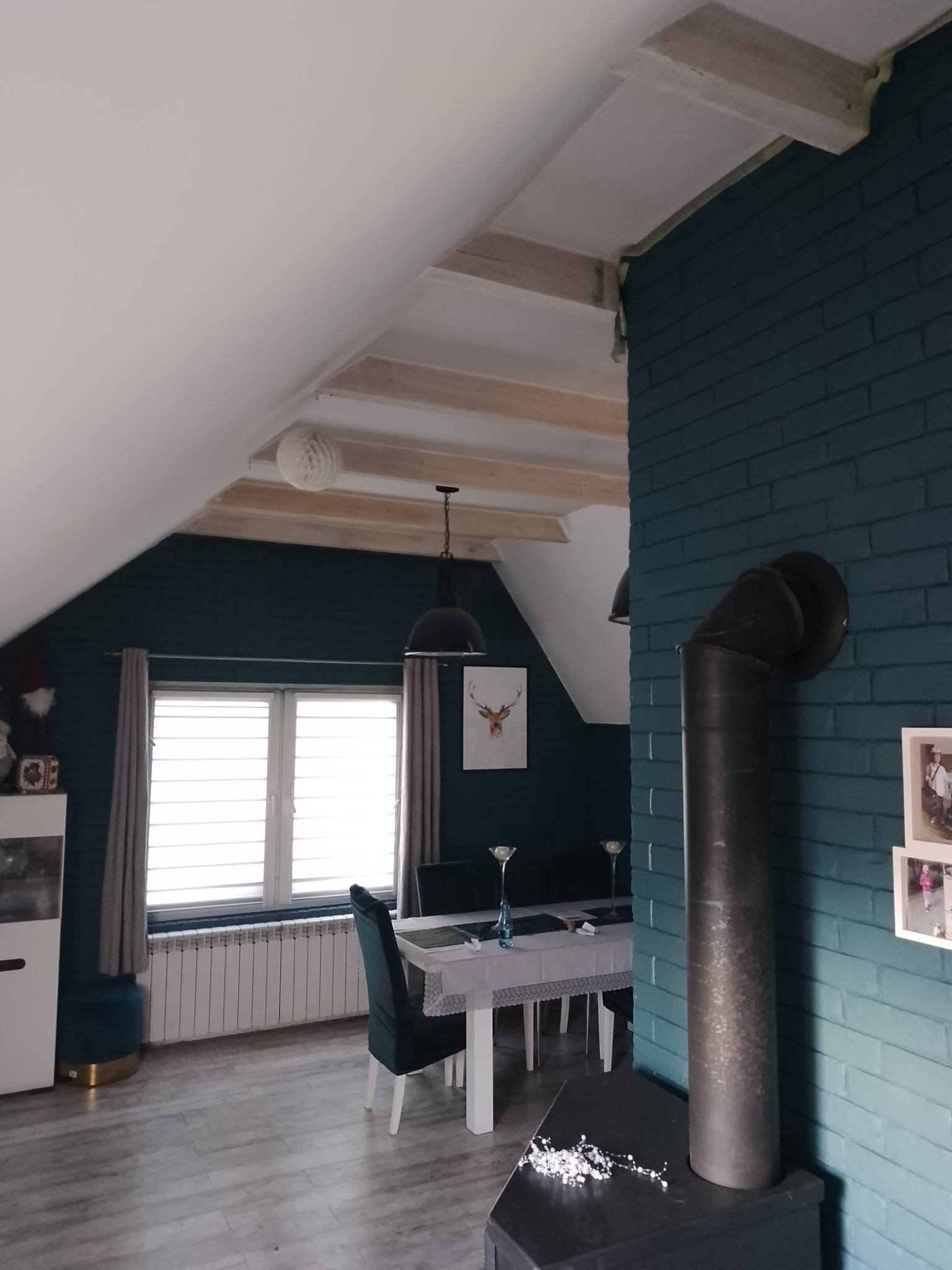 Poddasze mieszkalne z widocznymi belkami stropowymi, pomalowanymi cegłami na ścianie i kominkiem w rogu, oświetlone wiszącą lampą.