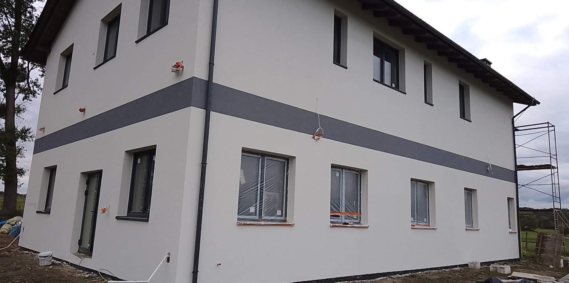 Dwukondygnacyjny dom z nową, jasną elewacją z poziomym pasem w kolorze szarym, okna zabezpieczone folią, widoczne rusztowanie z boku budynku.