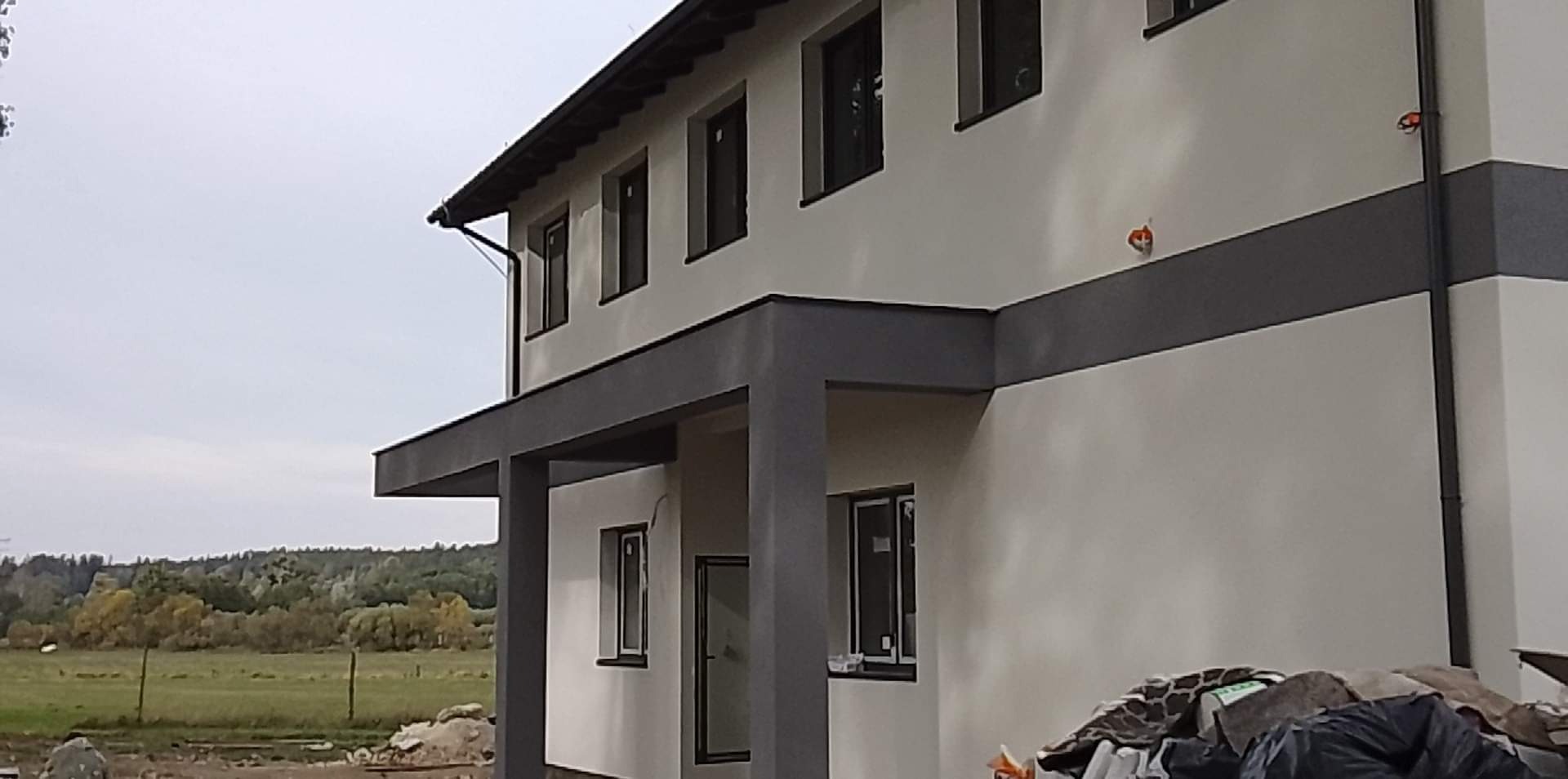 Dwukondygnacyjny dom z jasną elewacją i szarymi elementami dekoracyjnymi, widoczne okna z czarnymi ramami i teren budowy z przodu.