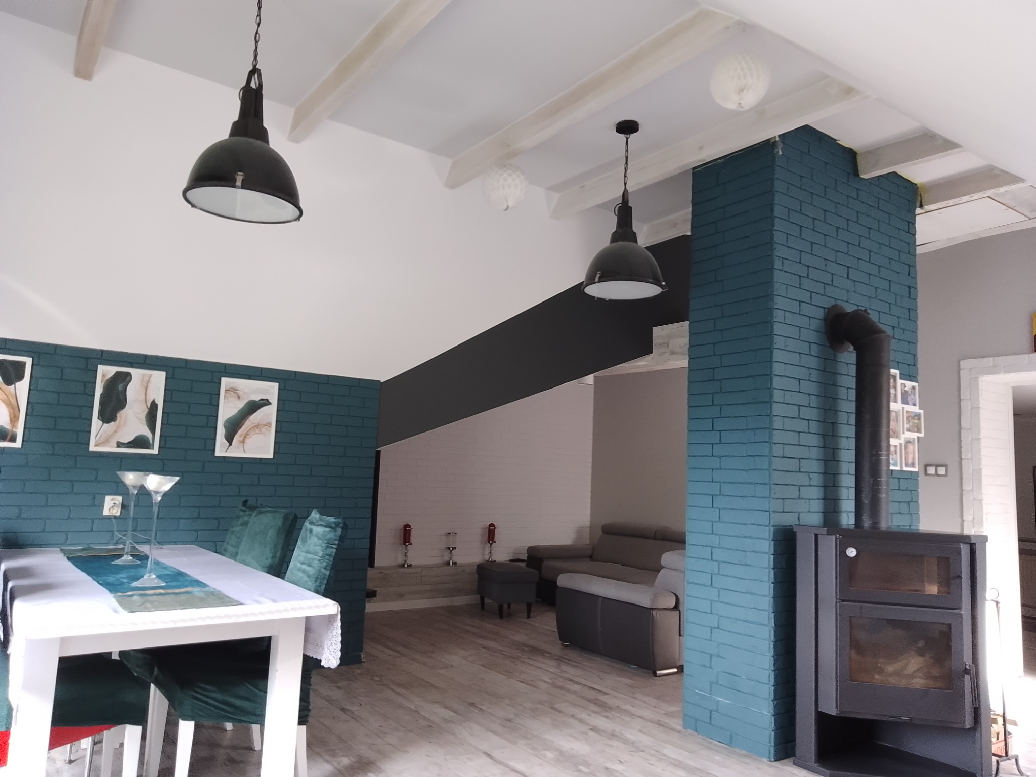 Salon z ceglaną ścianą pomalowaną na niebiesko, czarnym piecem kominkowym i białym stołem z zielonymi krzesłami. Dwa czarne, wiszące klosze oświetlają przestrzeń.