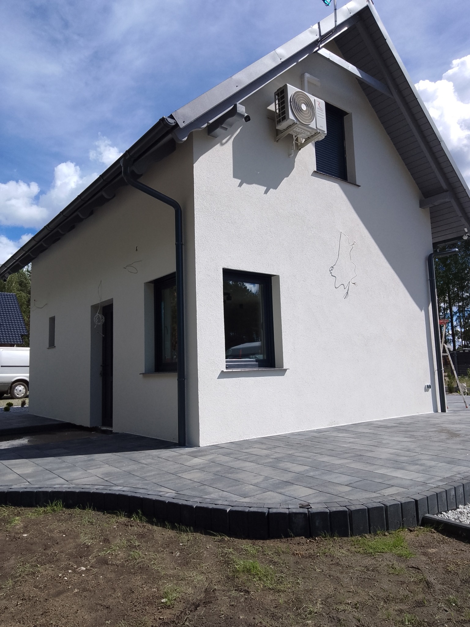 Wykończona elewacja domu jednorodzinnego z klimatyzatorem, widoczne ślady po instalacji elektrycznej, szara kostka brukowa wokół budynku.
