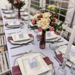 Elegancko nakryty stół na uroczystość w Czarnej Wsi, z białym obrusem, złotymi obramowaniami talerzy, różowymi serwetkami, szklankami, sztućcami, wazonami z kwiatami i małymi pudełkami z kokardkami...