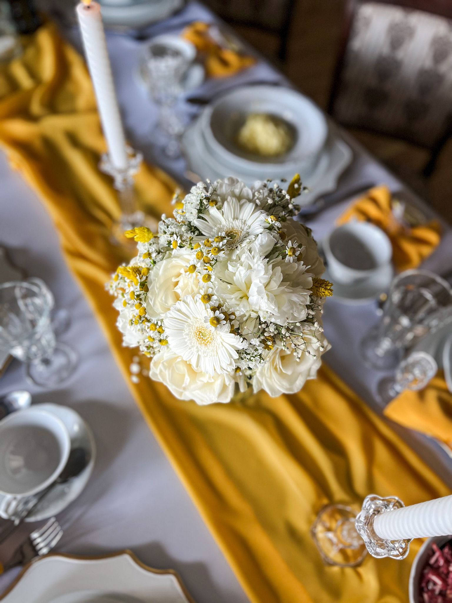 Elegancka dekoracja stołu z bukietem białych kwiatów z żółtymi akcentami, na złotym bieżniku, otoczona kryształowymi świecznikami i zastawą stołową w jasnych barwach.