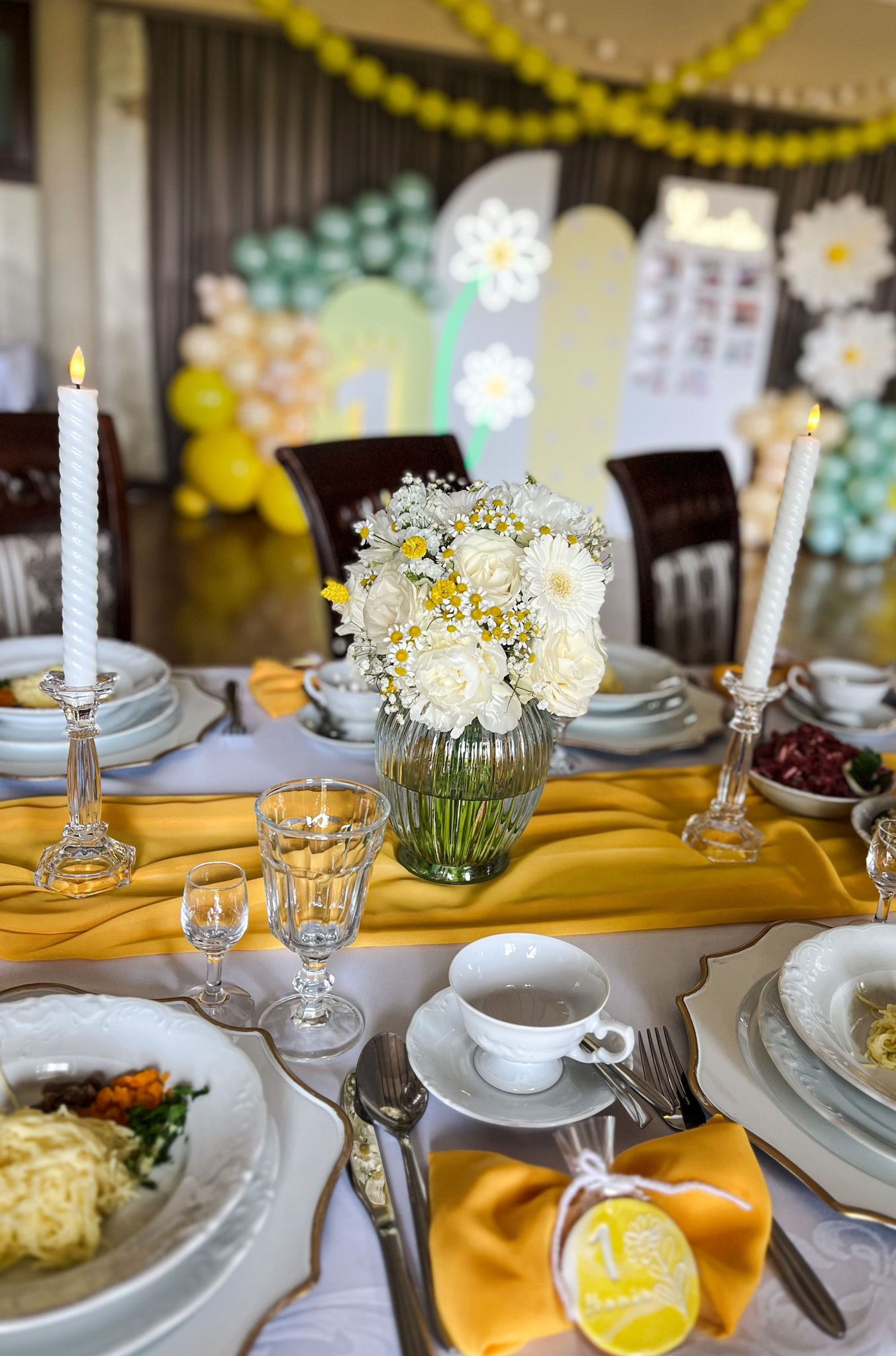 Elegancko nakryty stół z białymi i żółtymi kwiatami w szklanym wazonie, świecami w kryształowych świecznikach, białymi naczyniami ze złotym obramowaniem i żółtą dekoracją, w tle dekoracje balonowe.