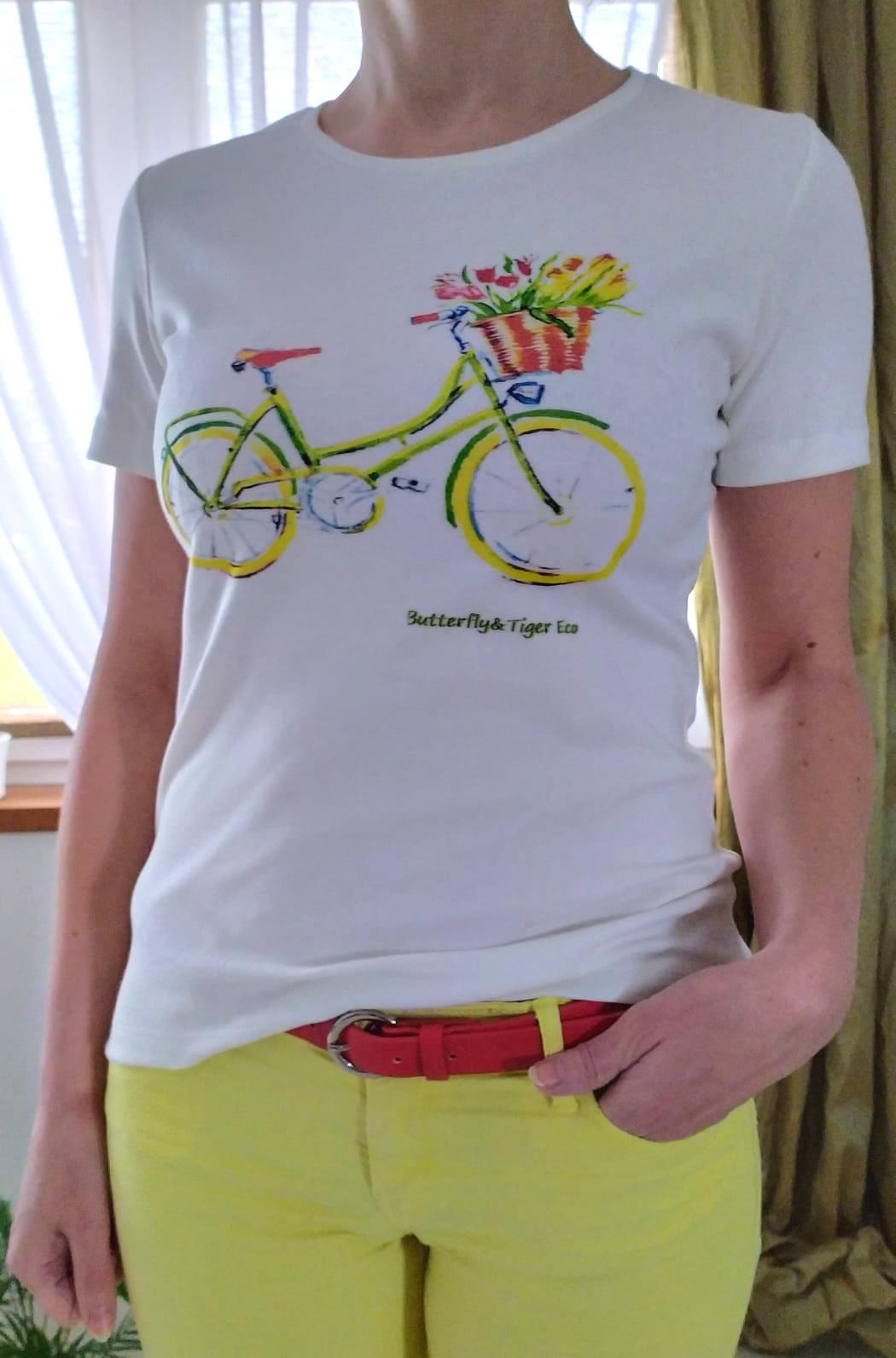 Kobieta w białej koszulce z nadrukiem roweru z kwiatami i napisem 'Butterfly&Tiger Eco', żółtych spodniach i czerwonym pasku.