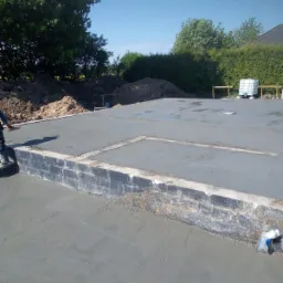 Wylana płyta fundamentowa z częściowo wymurowanym obrzeżem z bloczków, widoczne przygotowane podłoże i zbiornik na wodę w tle.