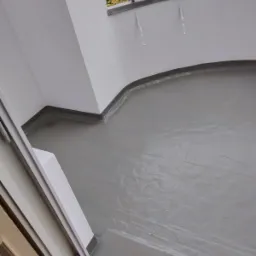 Szare, świeżo wylane podłoże na balkonie, z widocznymi detalami uszczelnienia przy ścianach i odpływem w rogu, widok z góry.