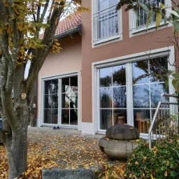 Dwukondygnacyjny dom z oknami PCV w białych ramach, brązową elewacją i balkonem z metalową balustradą, widok z zewnątrz w jesiennej scenerii.