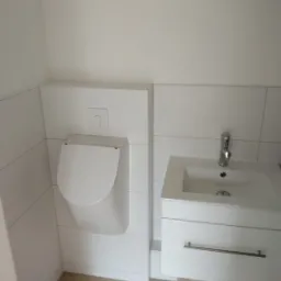 Biała, nowoczesna łazienka z wiszącym pisuarze i umywalką z szafką, wykończona białymi płytkami imitującymi drewno.