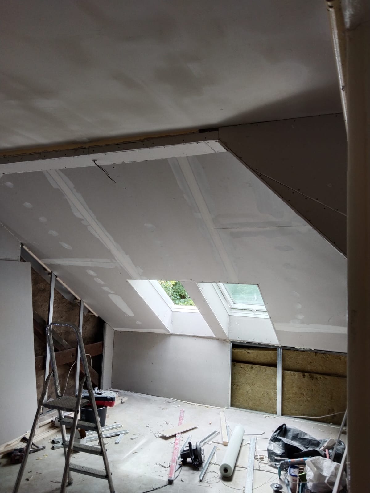Poddasze w trakcie adaptacji: widoczne płyty gipsowo-kartonowe na skośnym suficie z oknami dachowymi, częściowo ocieplone ściany, drabina i narzędzia na podłodze.