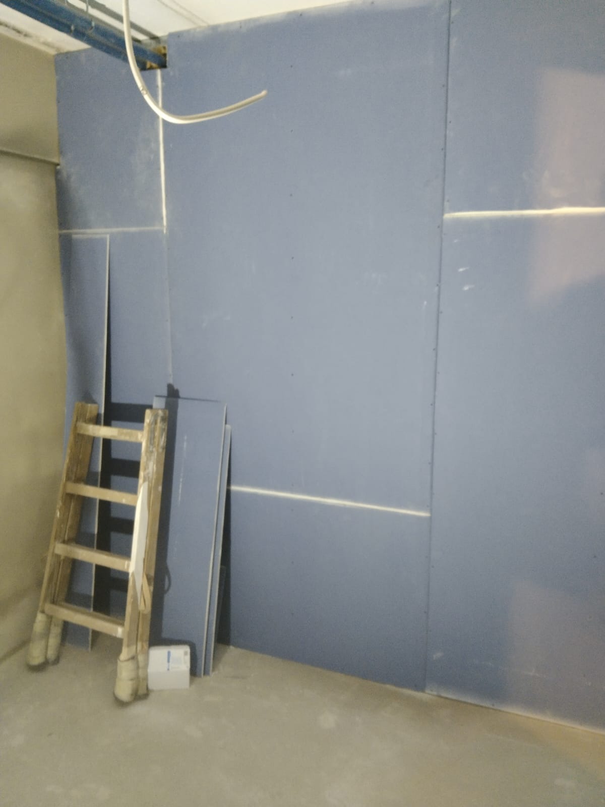 Montaż płyt gipsowo-kartonowych na ścianie, widoczna drewniana drabina oparta o ścianę oraz ułożone płyty g-k.