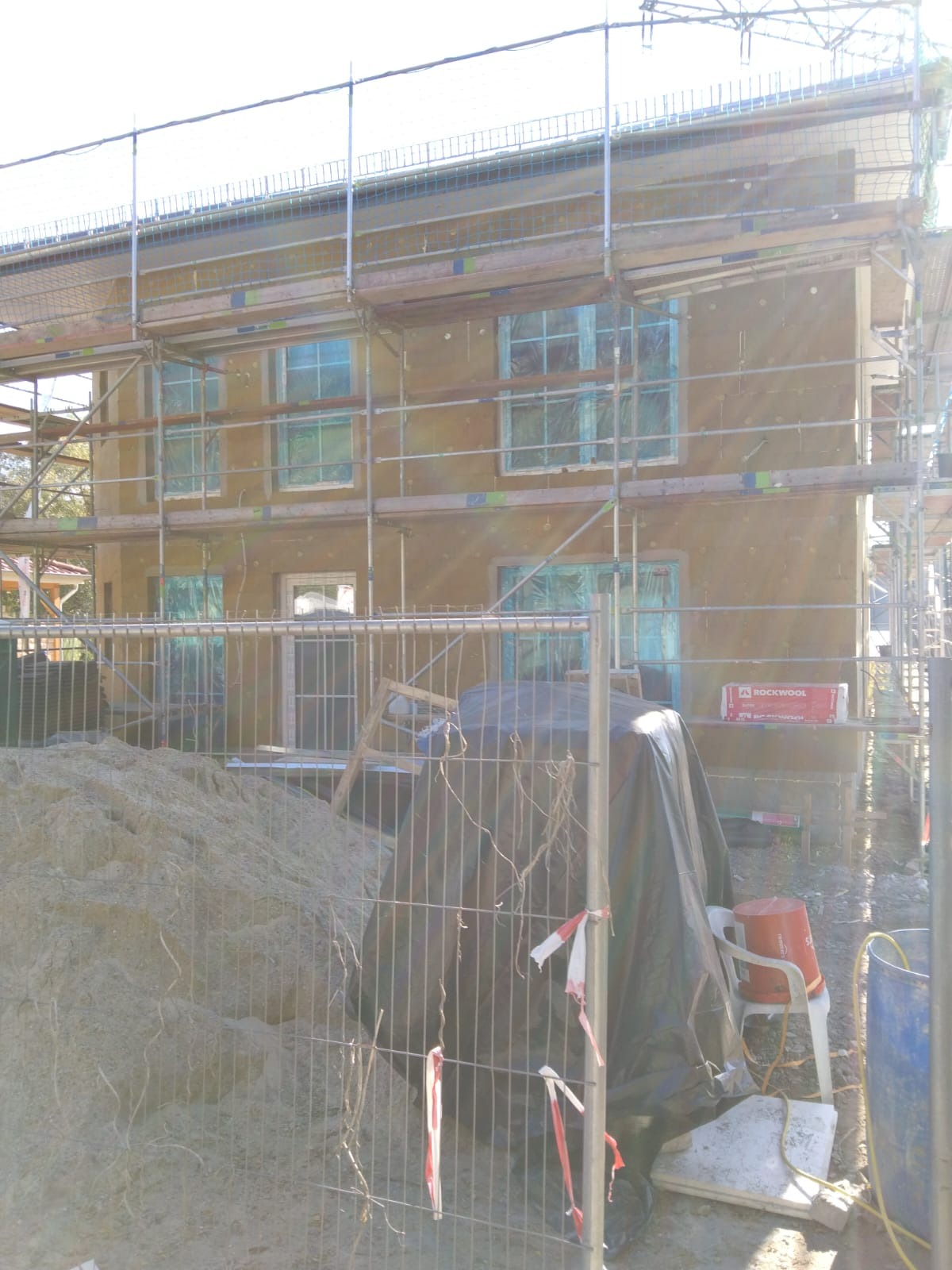 Dwupiętrowy dom w trakcie ocieplania, z rusztowaniem, zabezpieczonymi oknami folią, widoczny stos wełny mineralnej Rockwool i sterta piasku za ogrodzeniem.