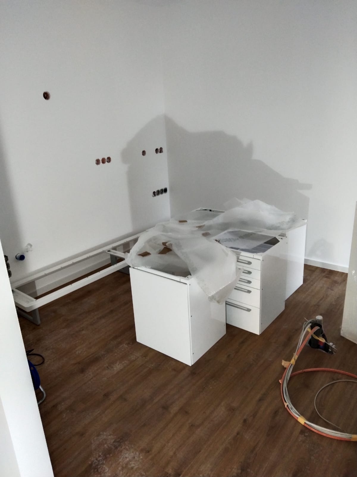 Montaż białych szafek kuchennych, częściowo jeszcze zabezpieczonych folią, na podłodze z paneli w trakcie prac wykończeniowych w pomieszczeniu z widocznymi puszkami elektrycznymi w ścianie.
