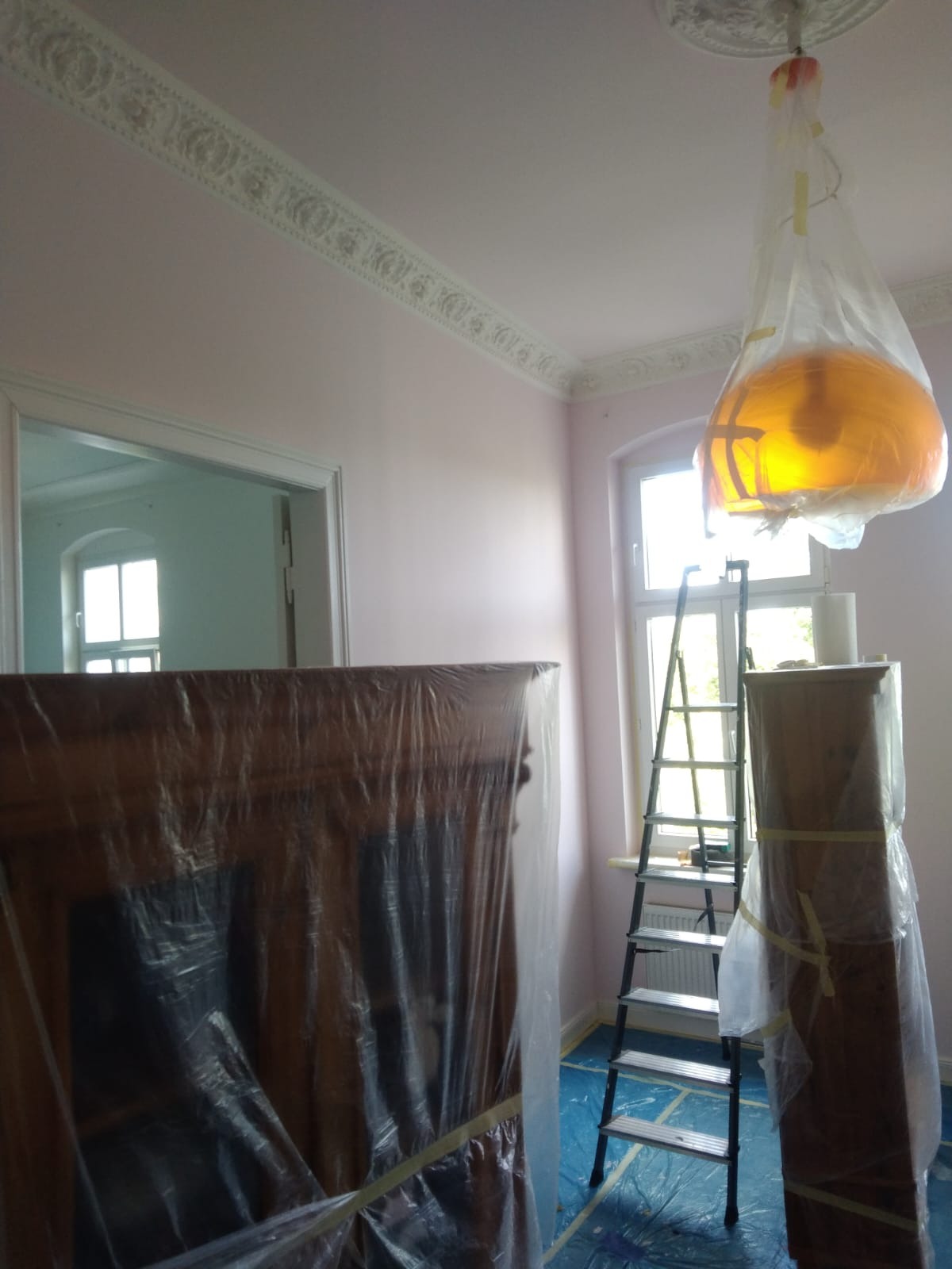 Pokój w trakcie malowania na różowo, meble zabezpieczone folią, drabina oparta o okno, lampa osłonięta workiem foliowym, widoczny ozdobny gzyms pod sufitem.