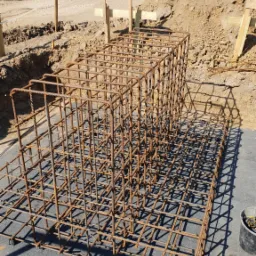 Klatka z prętów zbrojeniowych na szarej macie izolacyjnej pod fundament, w tle wykop pod fundament i pole uprawne w oddali.