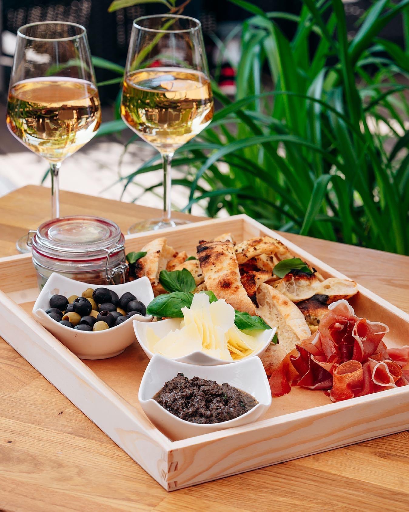 Drewniana taca z przekąskami: pieczywo, oliwki czarne i zielone w miseczkach, pesto, ser w płatkach, szynka parmeńska oraz dwa kieliszki białego wina na tle zieleni.