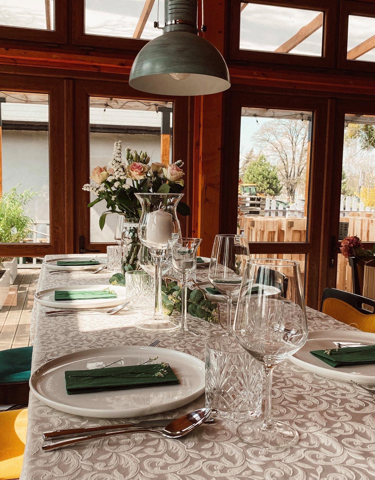 Elegancko nakryty stół z kwiatami i świecą w szklanym wazonie, zielone serwetki z drobnymi gałązkami na talerzach, widok przez okno na drzewa i plac budowy.