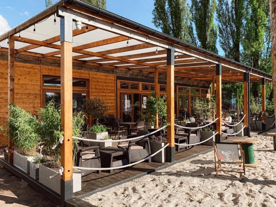 Drewniana restauracja z zadaszonym tarasem, rattanowe meble ogrodowe, rośliny w donicach, piasek na zewnątrz, tablica informacyjna z menu.