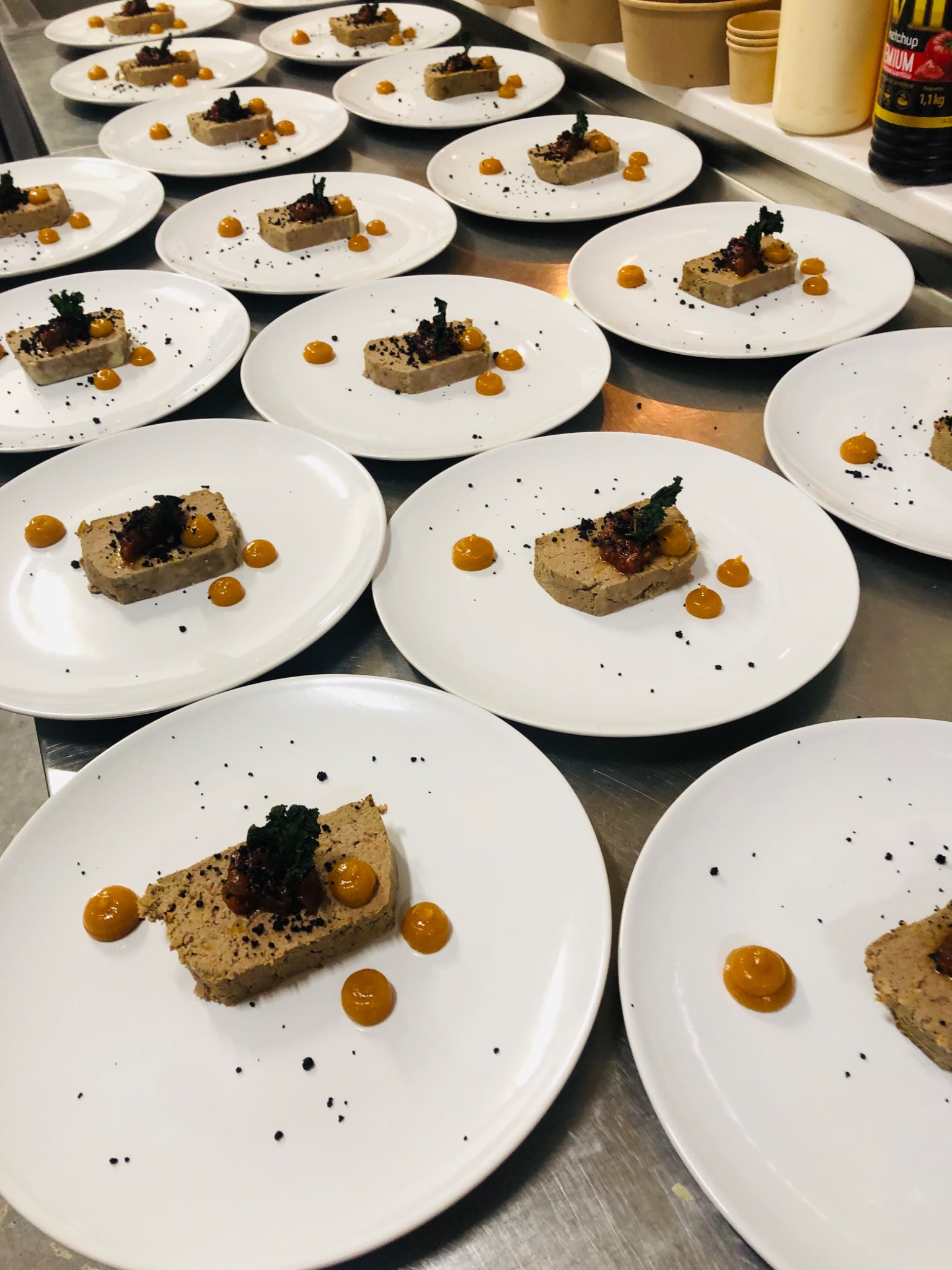 Przygotowane porcje pasztetu na białych talerzach z dekoracjami z kulek sosu, posypane czarnym pieprzem, ustawione na metalowym stole roboczym.