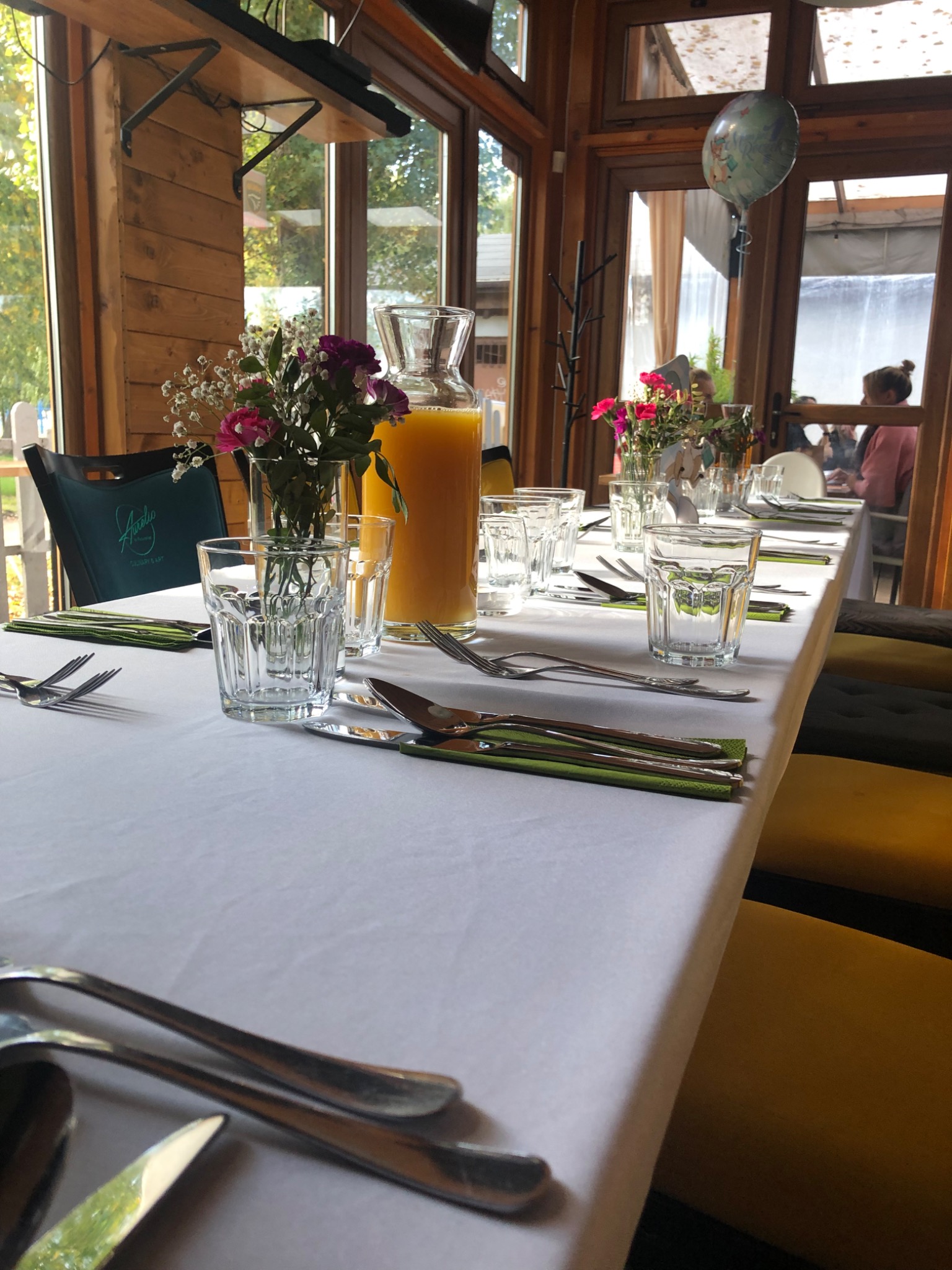 Ustawiony stół z białym obrusem, szklankami, sztućcami na zielonych serwetkach, wazonikami z kwiatami i karafką z sokiem pomarańczowym w jasnym wnętrzu z drewnianymi elementami.