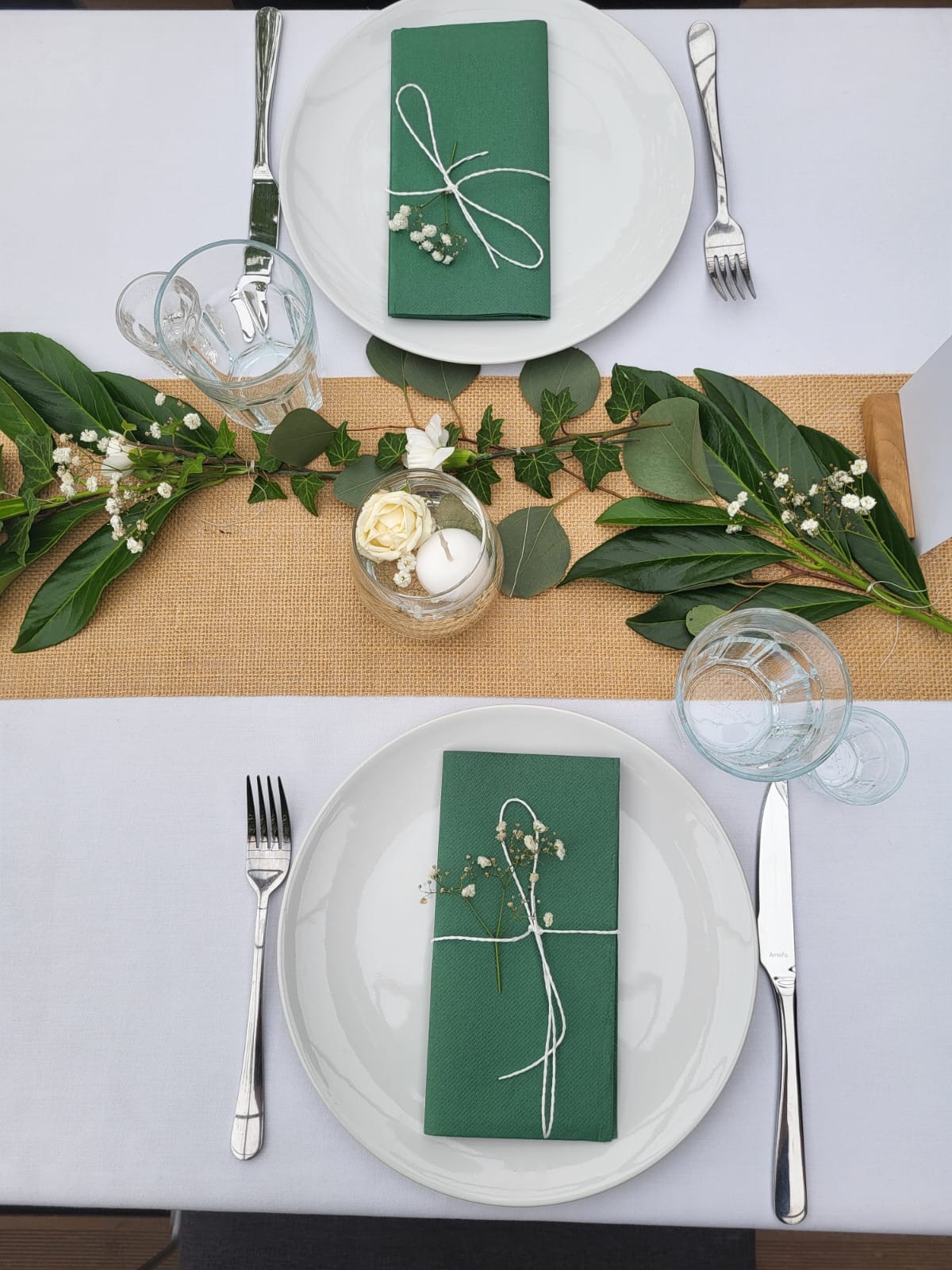 Elegancko nakryty stół z białym obrusem, jutowym bieżnikiem, zielonymi serwetkami przewiązanymi białym sznurkiem i drobnymi kwiatami, szklankami i sztućcami, dekorowany zielonymi liśćmi i kwiatami...