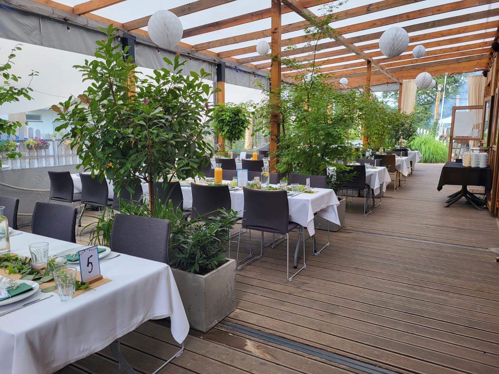 Uroczysta aranżacja stołów na drewnianym tarasie z roślinami w donicach, białymi obrusami, numerami stołów i dekoracjami z zieleni, oświetlona wiszącymi lampionami.