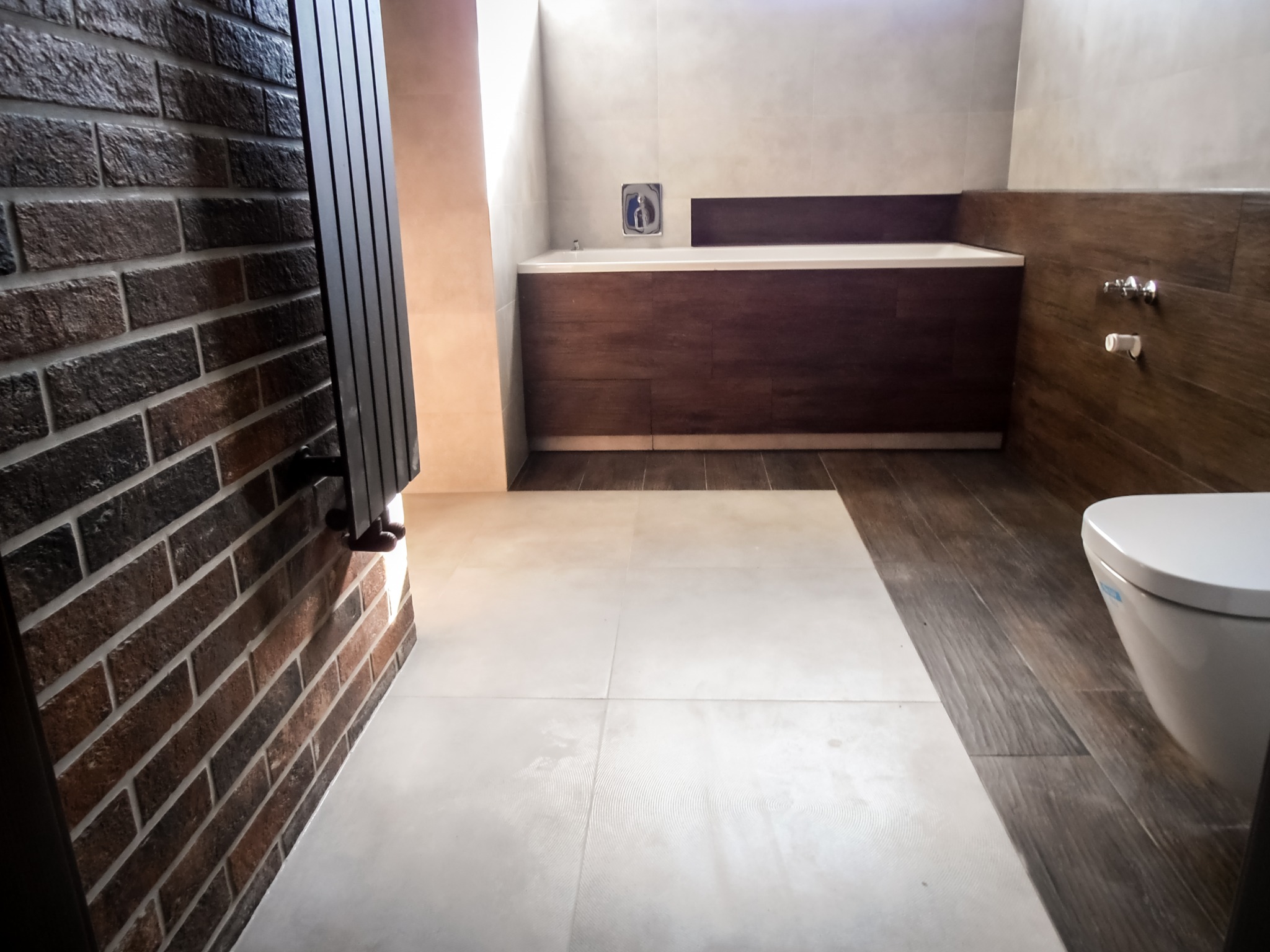 Nowoczesna łazienka z brązową cegłą na ścianie, grzejnikiem drabinkowym, wanną obudowaną drewnem i podłogą z płytek w odcieniach bieli i brązu.