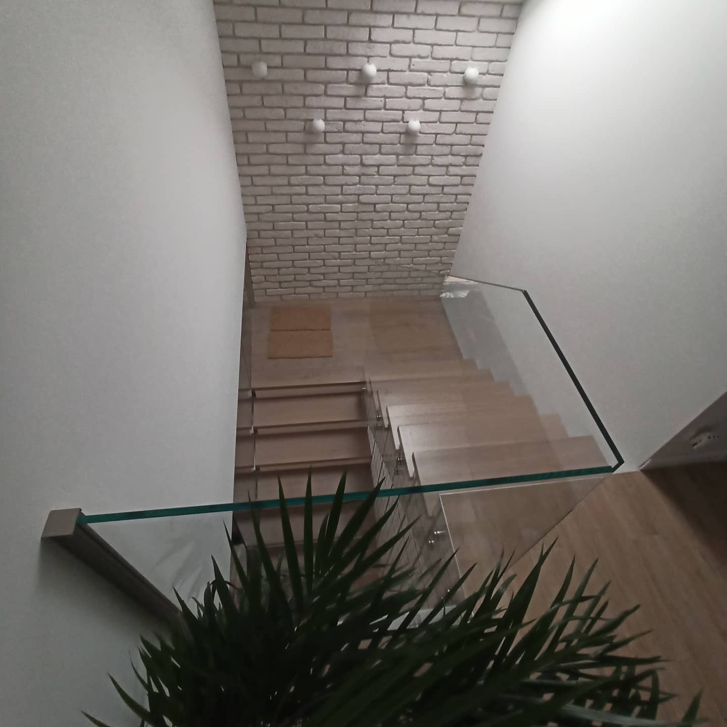 Schody drewniane z szklaną balustradą, widok z góry, z fragmentem ściany z białej cegły i rośliną doniczkową w rogu kadru.
