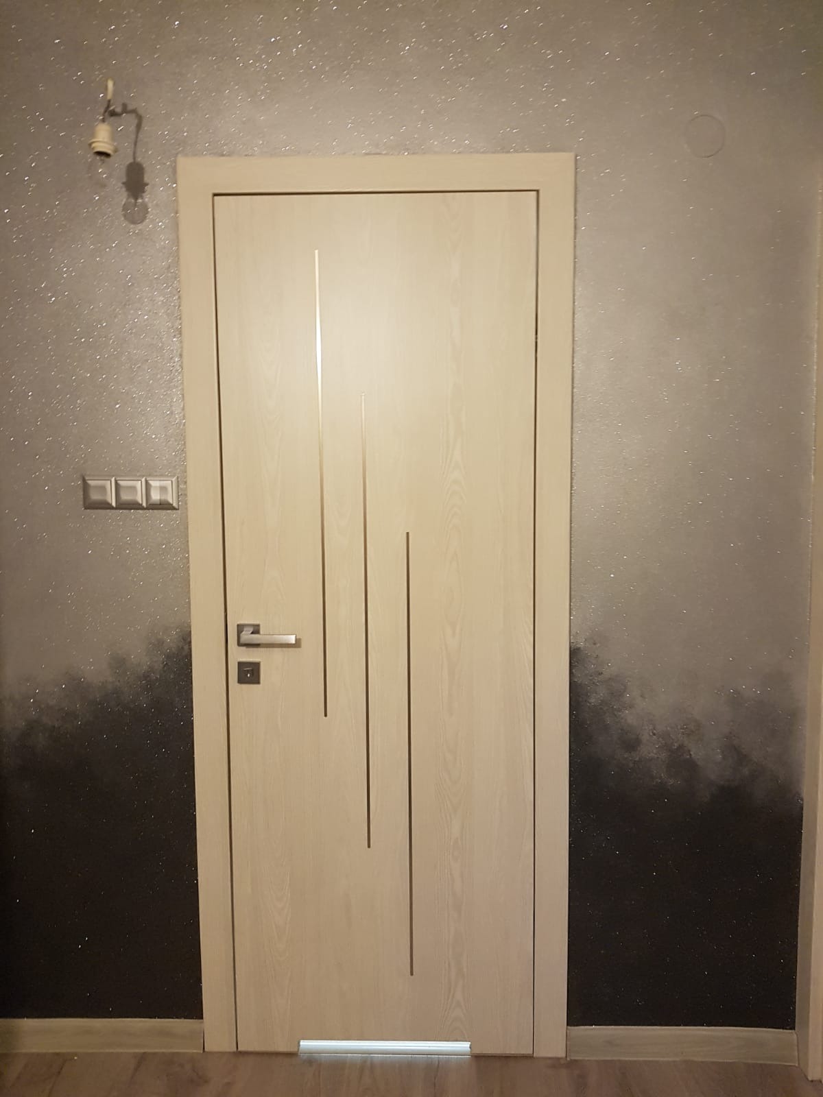 Jasne, drewnopodobne drzwi z trzema pionowymi wgłębieniami na tle ściany z efektem ombre przechodzącym od srebrnego brokatu u góry do czerni u dołu. Widoczny fragment parkietu i osprzęt elektryczny.