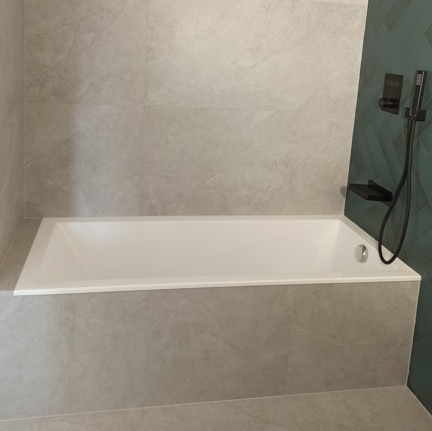 Biała wanna obudowana szarymi płytkami imitującymi kamień, z czarną armaturą prysznicową na ścianie wyłożonej zieloną jodełką.