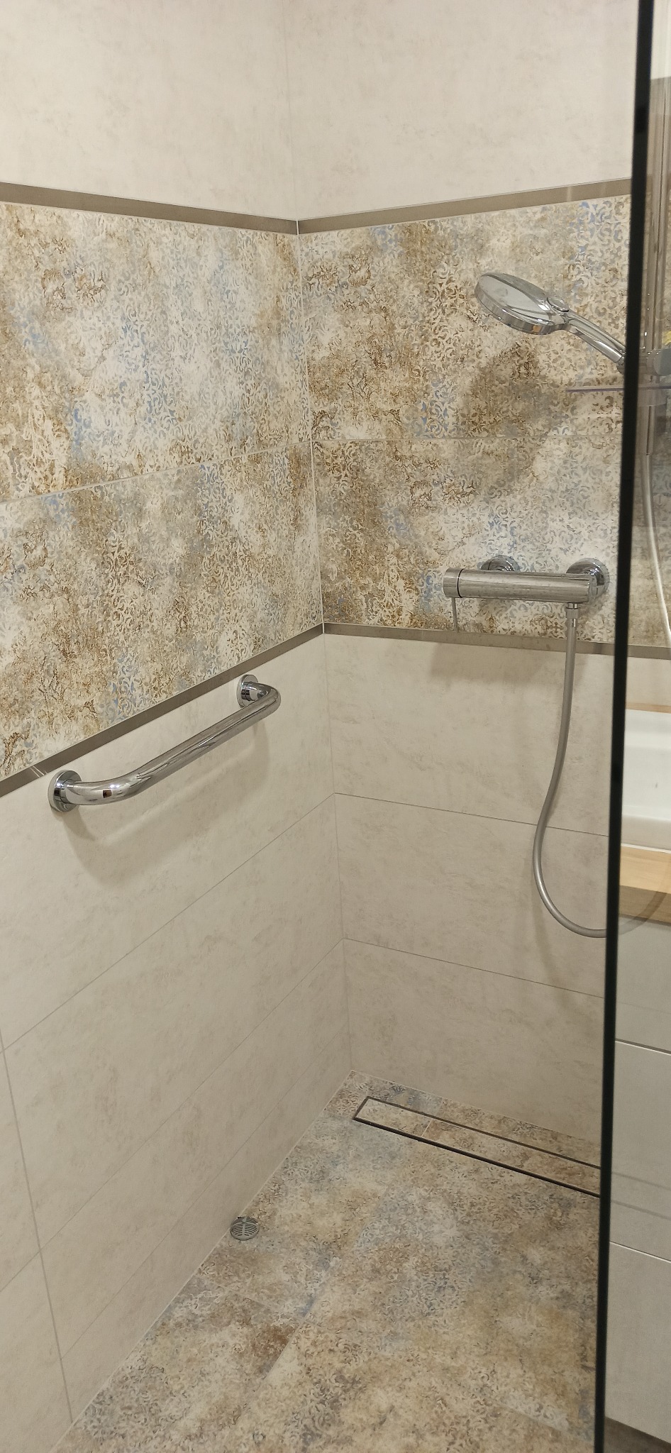 Wykończona kabina prysznicowa z beżowymi i brązowymi płytkami o wzorzystej fakturze, chromowanym uchwytem dla niepełnosprawnych i odpływem liniowym w podłodze.