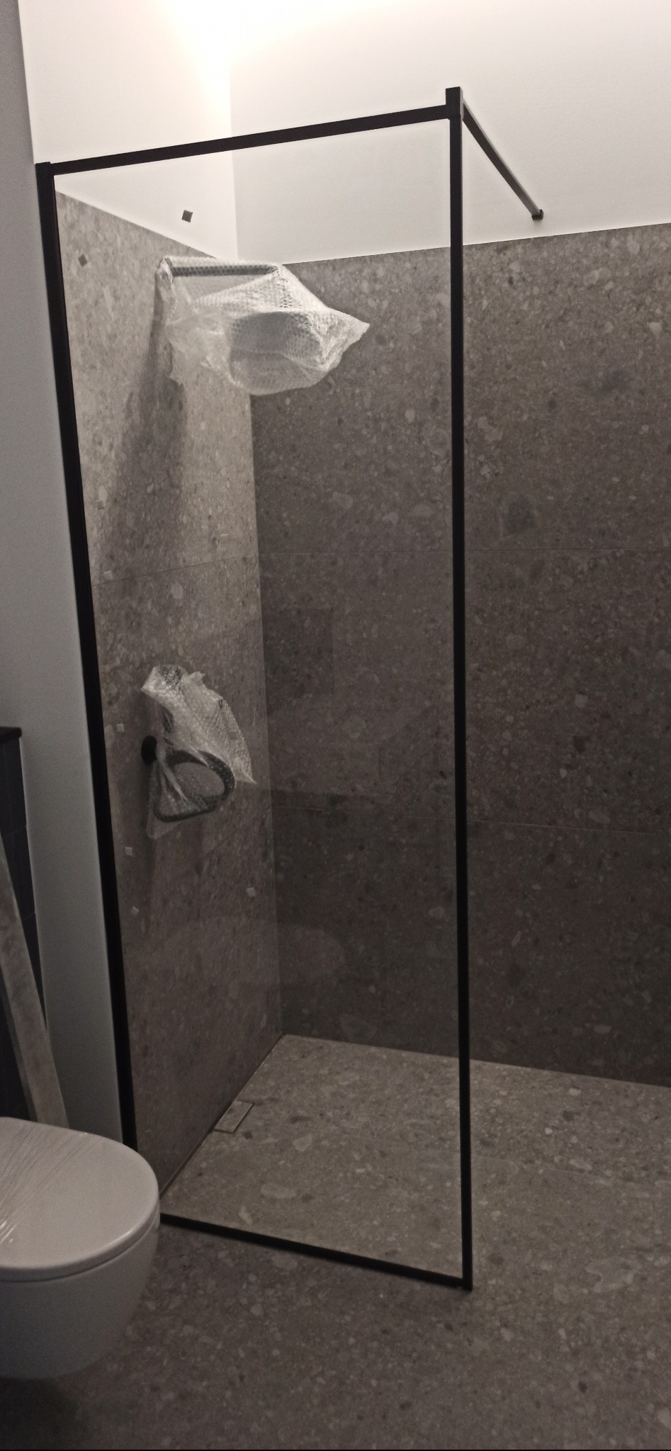 Szara łazienka z kabiną prysznicową typu walk-in z czarną ramą, armatura i deszczownica zabezpieczone folią, widoczna część toalety.