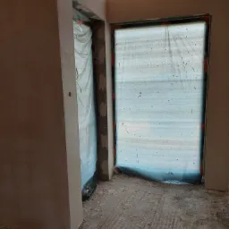 Surowe wnętrze w trakcie prac wykończeniowych: ściany pokryte świeżym tynkiem, okno i drzwi zabezpieczone folią malarską, widoczne drewniane elementy konstrukcji dachu, podłoga z resztkami...