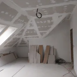 Poddasze w trakcie montażu płyt kartonowo-gipsowych na skosach dachu, widoczne szpachlowane łączenia, materiały budowlane na podłodze, okno dachowe w lewym górnym rogu.