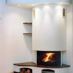 Nowoczesny narożny piec z paleniskiem obudowany białym, cylindrycznym murem z wnękami i drewnianymi półkami, płomienie w palenisku.