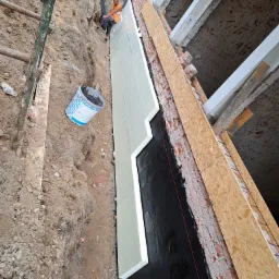 Fundament budynku w trakcie izolacji: widoczna czarna hydroizolacja, styropian, pracownik w odblaskowej kamizelce oraz wykop z ziemią i kubeł z zaprawą.