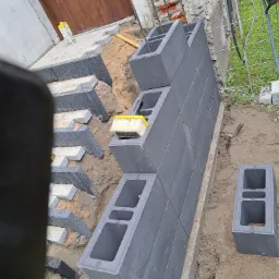 Budowa murku z szarych bloczków betonowych przy wejściu do domu, widoczny fragment kostki brukowej i narzędzia budowlane.