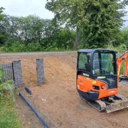 Pomarańczowa mini-koparka Kubota KX018-4 na placu budowy, widoczne fragmenty ogrodzenia z szarych słupków i podmurówki, w tle zieleń drzew i droga.