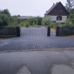 Nowa, ciemnoszara nawierzchnia z kostki brukowej prowadząca do domu z ogrodem, zakończona słupkami z ciemnego kamienia i drewnianym płotem, widok z asfaltowej drogi.